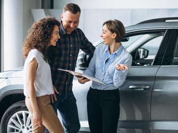 Ein Autoverkäufer bei Autoankauf Soest bespricht Details mit einem Paar und hält dabei ein Broschüre in der Hand, in einem modernen Autohaus, mit Fahrzeugen im Hintergrund.